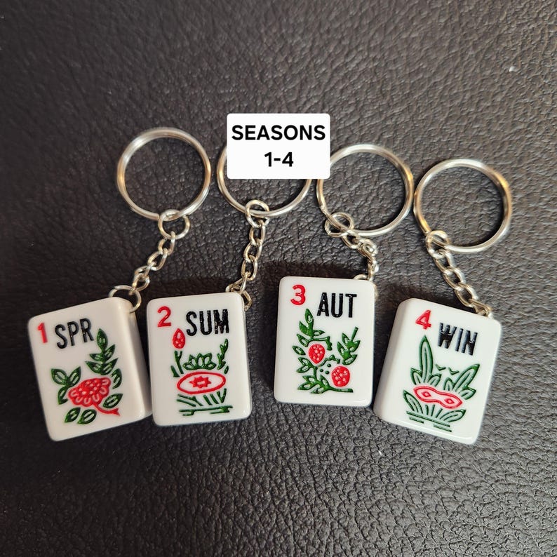 Puede incluir: Cuatro llaveros blancos con cadenas y anillas plateadas. Cada llavero tiene una baldosa blanca con un dise&ntilde;o verde y rojo y un n&uacute;mero del 1 al 4. Las baldosas dicen "1 SPR", "2 SUM", "3 AUT" y "4 WIN". El texto "SEASONS 1-4" est&aacute; impreso encima de los llaveros.