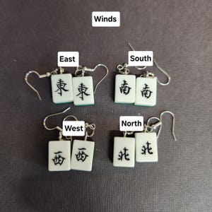 May include: Four pairs of earrings featuring white mahjong tiles with black Chinese characters representing the four winds: East, South, West, and North. The earrings are silver and have a hook closure.
