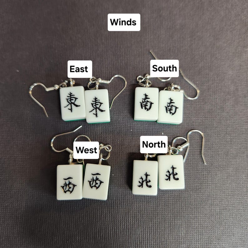 May include: Four pairs of earrings featuring white mahjong tiles with black Chinese characters representing the four winds: East, South, West, and North. The earrings are silver and have a hook closure.