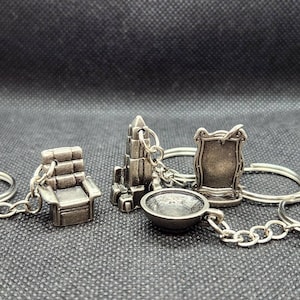 Könnte beinhalten: Ein Set aus fünf silbernen Schlüsselanhängern mit verschiedenen Designs, darunter ein Stuhl, ein Gebäude, ein Spiegel und eine Schüssel. Die Schlüsselanhänger sind an silbernen Schlüsselringen befestigt.