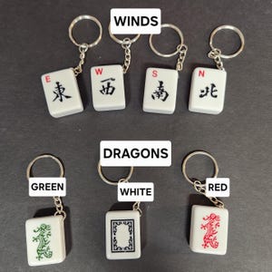 May include: Four white mahjong tile keychains with black characters and red lettering. The tiles represent the four winds: East, West, South, and North. Three white mahjong tile keychains with black and green, black and white, and black and red dragons. The tiles are labeled "Green", "White", and "Red".