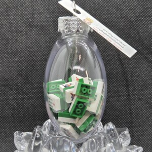 May include: A clear glass ornament filled with green and white toy blocks. The ornament has a silver cap and a tag that reads "www.etsy.com/shop/CrapBagBananaHammock". The ornament is resting on a clear, decorative base.