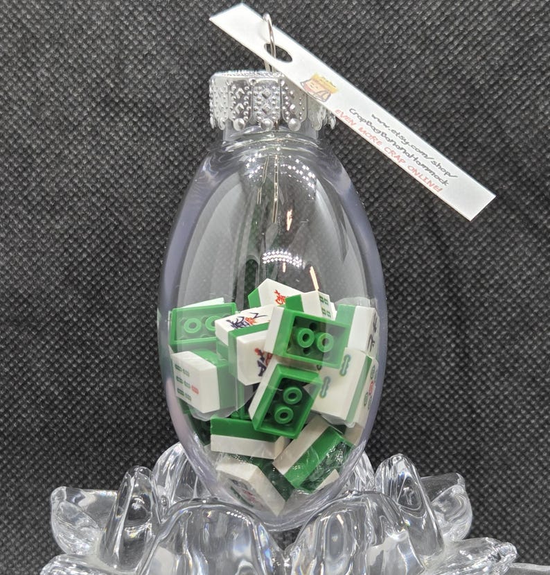 May include: A clear glass ornament filled with green and white toy blocks. The ornament has a silver cap and a tag that reads "www.etsy.com/shop/CrapBagBananaHammock". The ornament is resting on a clear, decorative base.