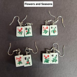 May include: A pair of earrings featuring four sets of mahjong tiles, each set with a different number and design. The tiles are white with green and red accents. The earrings are silver and have a hook closure.
