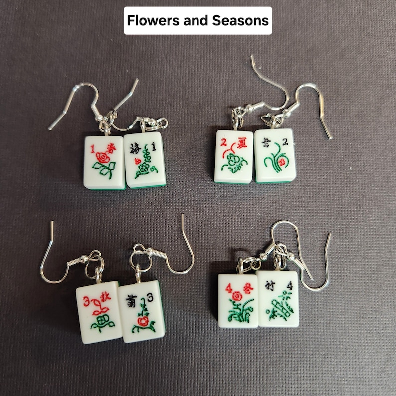 May include: A pair of earrings featuring four sets of mahjong tiles, each set with a different number and design. The tiles are white with green and red accents. The earrings are silver and have a hook closure.