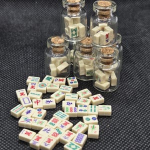 May include: A collection of miniature Mahjong tiles, some stored in small glass bottles with cork stoppers. The tiles are off-white with colorful symbols and characters. The background is a dark, textured surface.