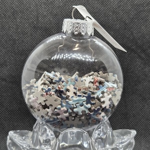 Puede incluir: Un adorno navideño de cristal transparente lleno de pequeñas piezas de rompecabezas en tonos azules, grises y rojos. El adorno tiene una tapa plateada y un lazo plateado para colgar. El adorno está sobre una base de cristal transparente.