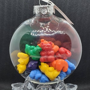 Può includere: Un addobbo natalizio in vetro trasparente pieno di orsetti gommosi colorati a forma di orsi e farfalle. L'addobbo è su un piedistallo in vetro trasparente.