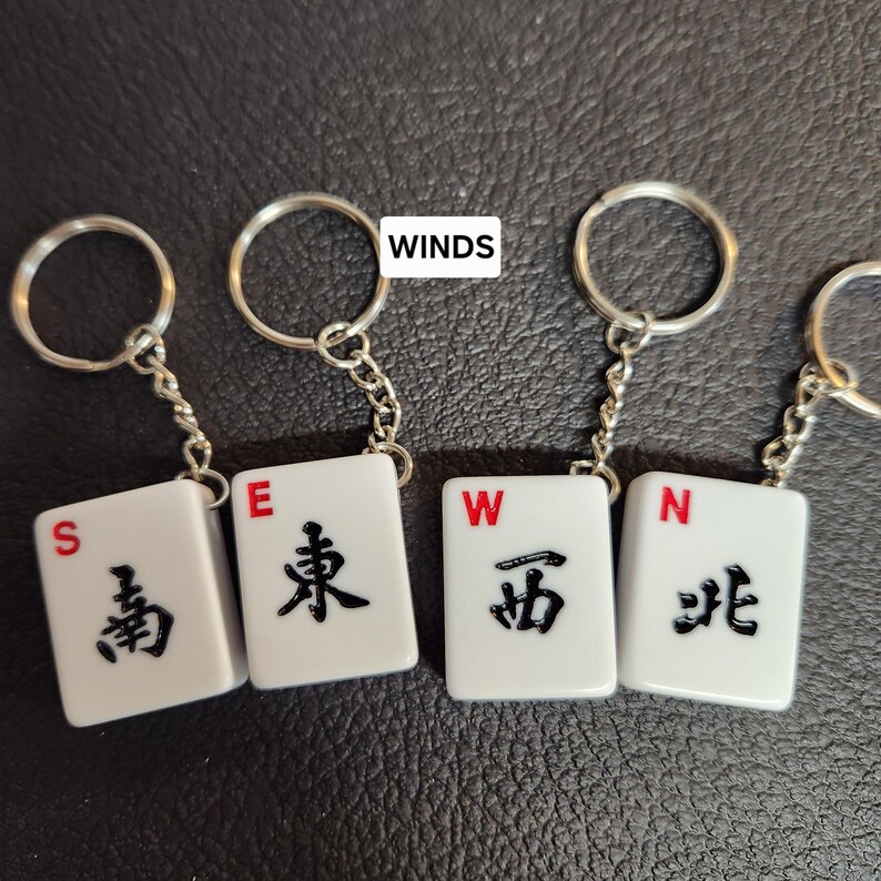 Puede incluir: Cuatro llaveros de fichas de mahjong blancas con caracteres chinos negros y letras rojas. Las fichas representan los cuatro vientos: Este, Sur, Oeste y Norte.