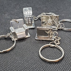 Pode incluir: Quatro chaveiros prateados com designs diferentes: uma cadeira, uma televisão, uma bandeja de comida e um teclado. Cada chaveiro tem um anel prateado.