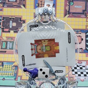 May include: A clear glass Christmas ornament with a white card inside. The card shows a floor plan of a room labeled "Library" with a knife and a game piece. The ornament is sitting on a game board with a checkered pattern.