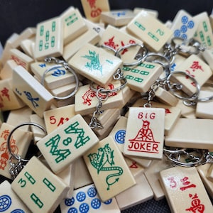 May include: A pile of off-white Mahjong tile keyrings. The tiles display symbols and numbers in green, red, and blue. One tile reads "BIG JOKER" in red. The keyrings are attached with silver-coloured metal rings.