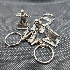 May include: A collection of silver-colored keychains featuring various designs. The keychains include a figure of a person, an owl, a pair of shoes, and a flag. Each keychain has a silver ring for attaching keys.