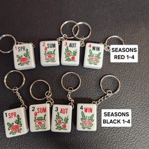 May include: Four white tile keychains with red and green designs. The tiles are labeled with numbers 1-4 and the words "SPR", "SUM", "AUT", and "WIN". The keychains are labeled "SEASONS RED 1-4" and "SEASONS BLACK 1-4".