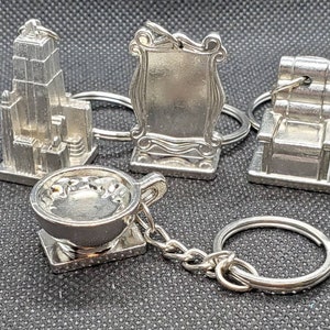 Könnte beinhalten: Eine Sammlung silberfarbener Schlüsselanhänger. Die Schlüsselanhänger zeigen ein Empire State Building, ein gerahmtes Schild, einen Stuhl und eine Teetasse mit Untertasse. Die Schlüsselanhänger sind aus Metall und an silbernen Schlüsselringen befestigt.