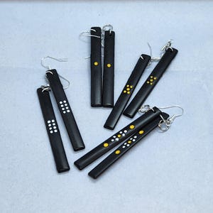 Peut inclure: Un ensemble de six boucles d'oreilles pendantes noires avec des points blancs et dorés. Les boucles d'oreilles sont longues et fines, et elles sont parfaites pour ajouter une touche de style à n'importe quelle tenue.