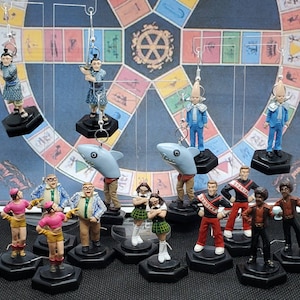 May include: A collection of 12 colorful, whimsical figurines, including a shark, a mermaid, and a variety of people. The figurines are displayed on a game board with a circular design.