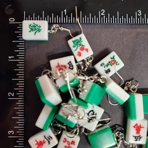 May include: A collection of green and white mahjong tile earrings with silver hooks. The tiles feature Chinese characters and symbols.