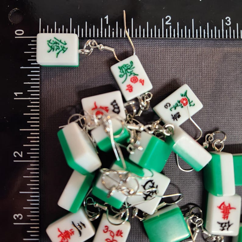 May include: A collection of green and white mahjong tile earrings with silver hooks. The tiles feature Chinese characters and symbols.