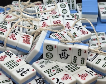 Op de afbeelding: Een verzameling witte en blauwe Mahjong-tegels met rode en zwarte symbolen en karakters. De tegels zijn aan elkaar geregen met een natuurlijk gekleurd koord. De afbeelding toont een verscheidenheid aan tegels, waaronder die met cirkels, bamboe en Chinese karakters.