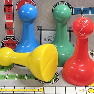 Puede incluir: Cuatro coloridas fichas de juego en azul, verde, rojo y amarillo están dispuestas sobre un juego de mesa. Las piezas tienen una parte superior redondeada y una base larga y cónica. El tablero del juego tiene el texto "HOME", "SAFETY ZONE" y "START".