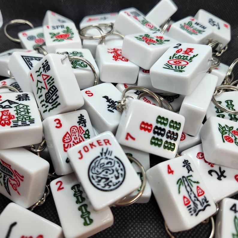 Puede incluir: Una colecci&oacute;n de llaveros de fichas de mahjong blancas con marcas negras y rojas. Las fichas presentan varios n&uacute;meros, s&iacute;mbolos y caracteres, incluyendo "JOKER", "WIN", "1福", "2", "3", "4", "7", y "S".