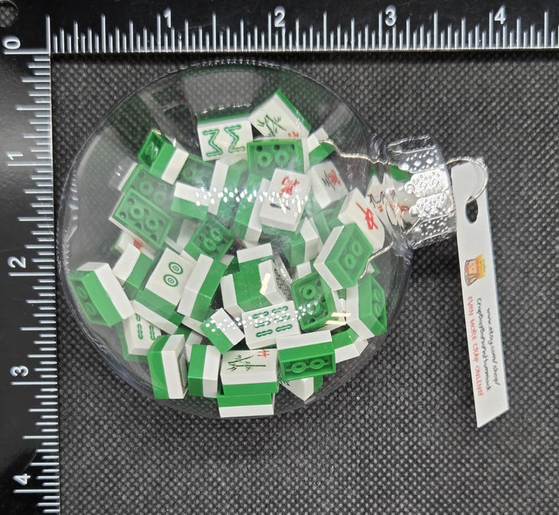 May include: A clear glass Christmas bauble filled with miniature green and white Mahjong tiles. The bauble has a silver cap and a small tag attached. The tiles are scattered inside the glass sphere.