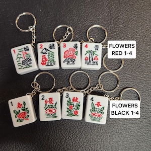 Puede incluir: Ocho llaveros de fichas de mahjong blancas con caracteres y s&iacute;mbolos chinos rojos y verdes. Las fichas est&aacute;n numeradas del 1 al 4 y est&aacute;n etiquetadas como "FLOWERS RED 1-4" y "FLOWERS BLACK 1-4".