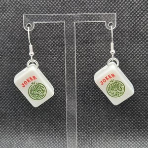May include: A pair of white earrings with a rectangular charm featuring the word "JOKER" in red and a green design. The earrings have silver-coloured hooks and are displayed against a dark background.
