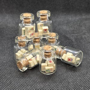 Peut inclure: Petites bouteilles en verre remplies de tuiles de Mahjong miniatures. Chaque bouteille a un bouchon en liège. Les tuiles sont blanc cassé avec des caractères et symboles chinois en rouge, vert et bleu. Les bouteilles sont disposées sur une surface sombre.