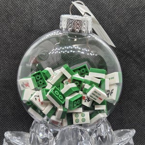 May include: A clear glass bauble filled with green and white Mahjong tiles. The bauble has a silver cap and a metal loop for hanging. The tiles feature various designs and symbols.