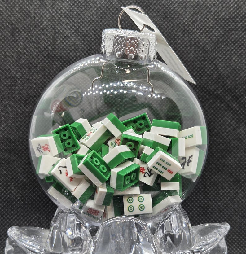 May include: A clear glass bauble filled with green and white Mahjong tiles. The bauble has a silver cap and a metal loop for hanging. The tiles feature various designs and symbols.