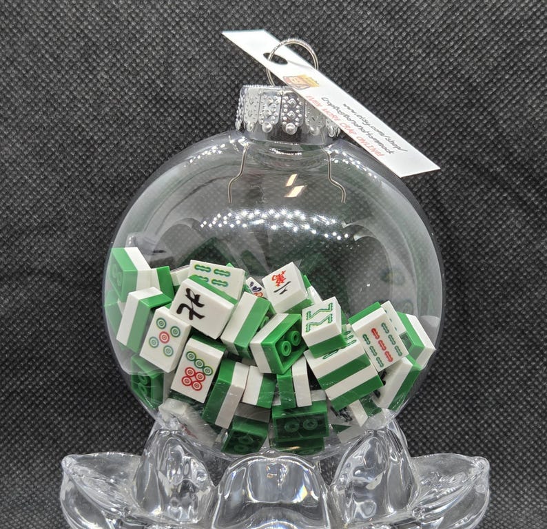 May include: A clear glass Christmas bauble filled with miniature green and white Mahjong tiles. The bauble has a silver cap and a small tag attached. The bauble is resting on a clear, decorative base.
