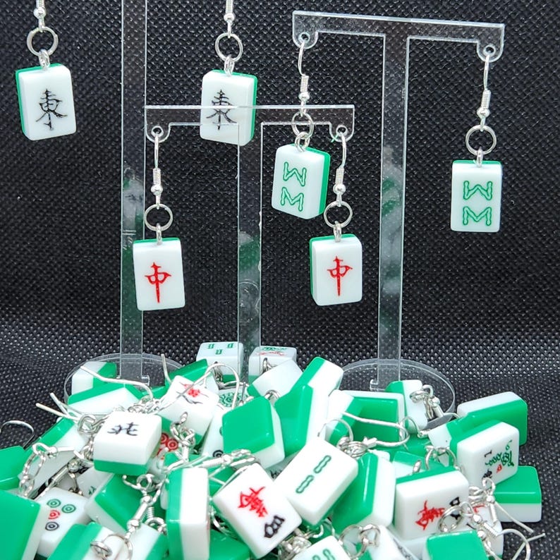 May include: A collection of green and white mahjong tile earrings. The tiles feature Chinese characters and symbols. The earrings are displayed on a clear acrylic stand.