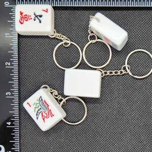 May include: Four white ceramic mahjong tiles with black and red Chinese characters and symbols attached to silver keychains. The tiles are connected by a silver chain with silver rings.