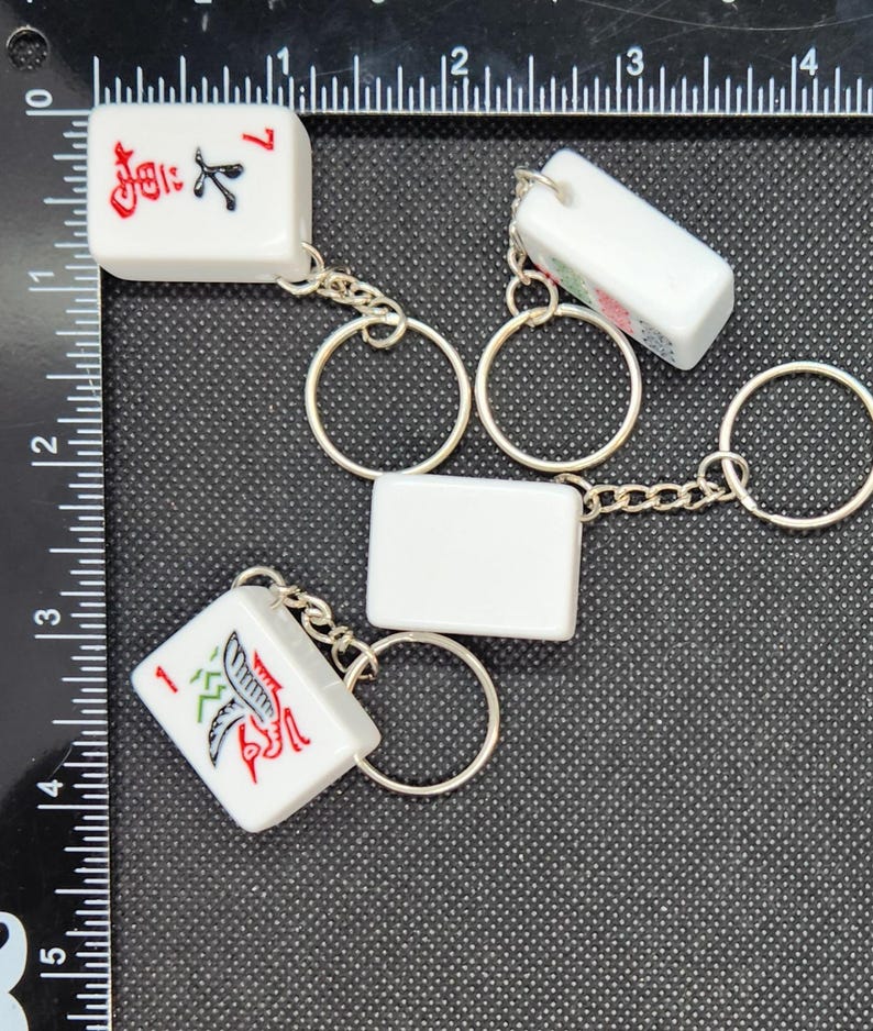 May include: Four white ceramic mahjong tiles with black and red Chinese characters and symbols attached to silver keychains. The tiles are connected by a silver chain with silver rings.