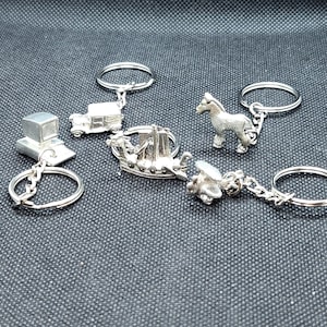 Puede incluir: Una colección de llaveros plateados con diseños en miniatura de un coche, un caballo, un barco, un avión y una caja sobre una balanza. Cada llavero tiene un anillo y una cadena plateados.