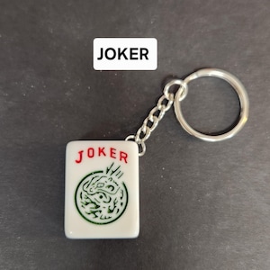 May include: A white keychain with a red "JOKER" and a green dragon design. The keychain is attached to a silver chain and a silver ring.