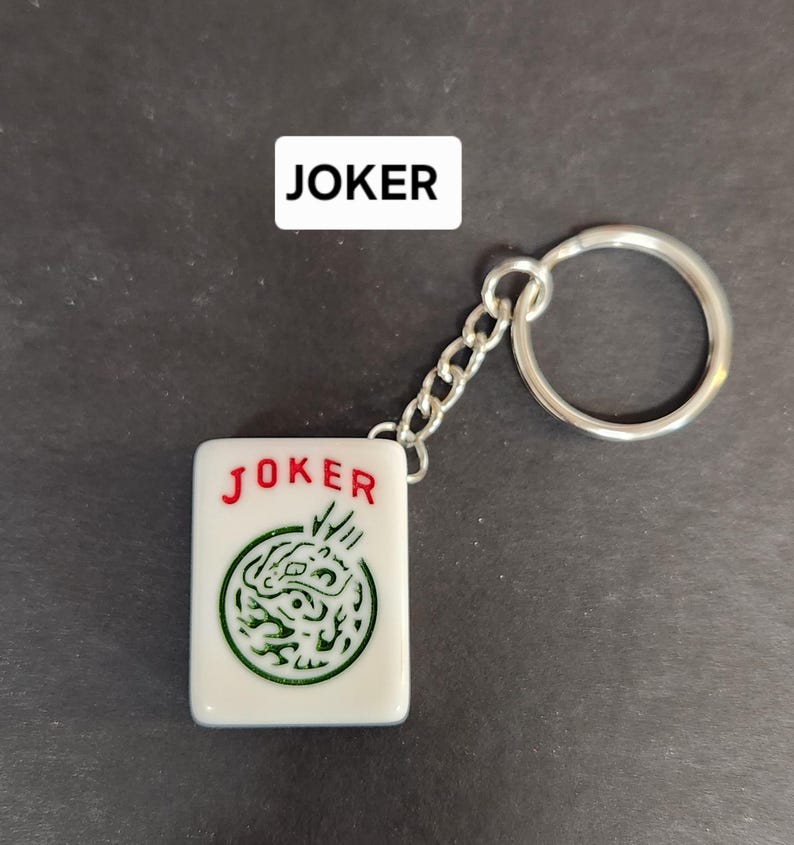 May include: A white keychain with a red "JOKER" and a green dragon design. The keychain is attached to a silver chain and a silver ring.