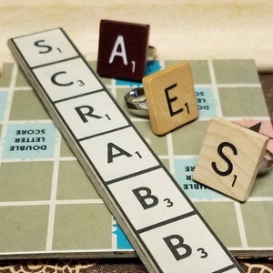 May include: Five Scrabble tile rings with the letters S, C, R, A, and E. The rings are made of wood and have a brown or beige finish. The tiles are arranged in a row on a Scrabble game board.
