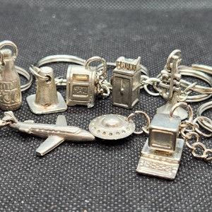 May include: A collection of silver-toned keychain charms, including a bottle of soda, a mail box, a phone booth, a trumpet, a UFO, a plane, and a computer.