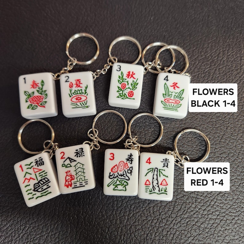 May include: Set of eight white ceramic mahjong tile keychains. The tiles are numbered 1-4 and feature Chinese characters and symbols. The tiles are divided into two sets: black with red characters and red with black characters. The text "FLOWERS BLACK 1-4" and "FLOWERS RED 1-4" is printed below each set.