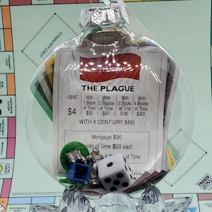 Puede incluir: Adorno navideño de cristal transparente lleno de piezas del juego Monopoly y una tarjeta de propiedad con la etiqueta "THE PLAGUE". El adorno incluye una tapa plateada y un lazo de plástico transparente para colgar. Contiene un dado, una casa azul y una pieza plateada.