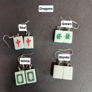 May include: Four pairs of earrings featuring mahjong tiles. The tiles are white with green and red accents. The tiles are labeled "Dragons", "Green", "Red", and "White".