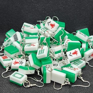 May include: A collection of green and white mahjong tile earrings. Each earring features a different mahjong tile with Chinese characters and symbols. The earrings are made of metal and have silver hooks.