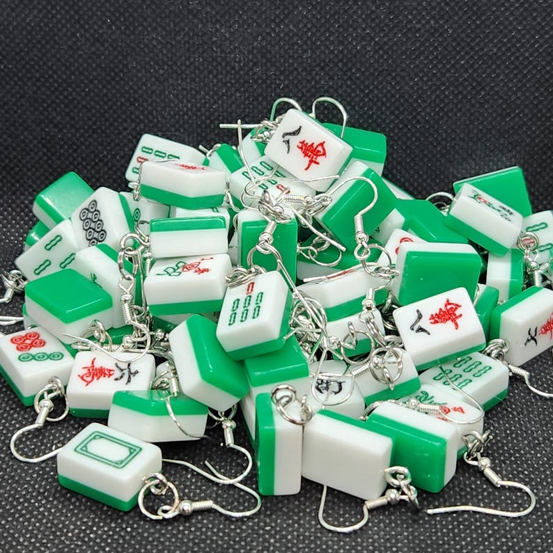 May include: A collection of green and white mahjong tile earrings. Each earring features a different mahjong tile with Chinese characters and symbols. The earrings are made of metal and have silver hooks.