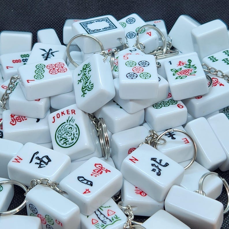 May include: A collection of white mahjong tile keychains with various Chinese characters and symbols. The tiles are decorated with red, green, and black details. The keychains are attached to silver key rings.