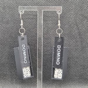 Op de afbeelding: Een paar zwarte domino-oorbellen. Elke oorbel heeft een kleine, rechthoekige doos met het woord "DOMINO" in witte letters. In elke doos zitten witte dominostenen met zwarte stippen. De oorbellen hangen aan zilverkleurige haken.