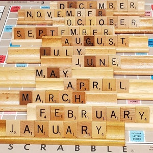 May include: A Scrabble board displays the months of the year spelled out with wooden letter tiles. The months are arranged in descending order, from December at the top to January at the bottom. The board's grid and score areas are visible.