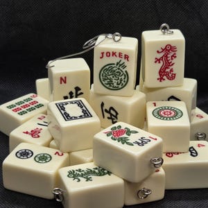 Op de afbeelding: Een verzameling ivoorkleurige Mahjong-tegels met diverse symbolen en karakters in rood, groen en zwart. Sommige tegels hebben metalen lussen. De tegels tonen afbeeldingen van draken, bloemen en het woord "JOKER".